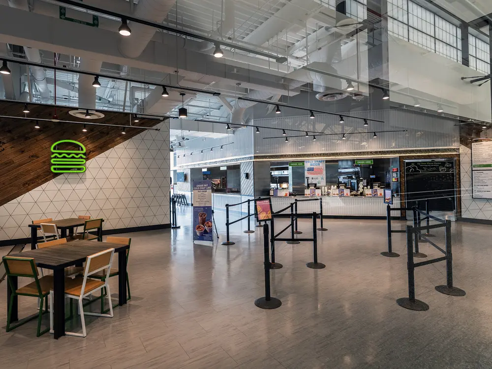 An empty fast food cafe. On the left is a table under the green Shake Shack logo. On the right is a cordoned rope leading up to the counter. 