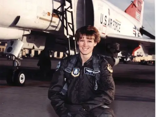A woman posing next to an aircraft in a military flight suit.