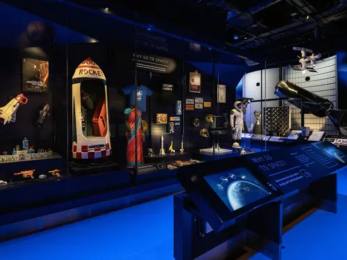 A display case featuring a variety of objects. From left to right you can see model spacecraft, from toy size to 5 feet tall, a sari, a t-shirt, and a spacesuit. In front of it is a long text panel. 