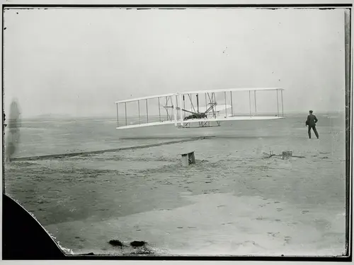 A biplane just barely above the ground with a person standing to the right of it.