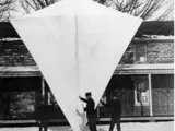 A giant four sided kite. Several men stand in front of the kite, and it is more than twice as tall as they are. 