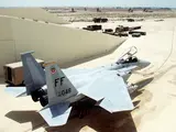A U.S. Air Force F-15 Eagle fighter jet parked on a tarmac, with various military vehicles and equipment visible in the background.