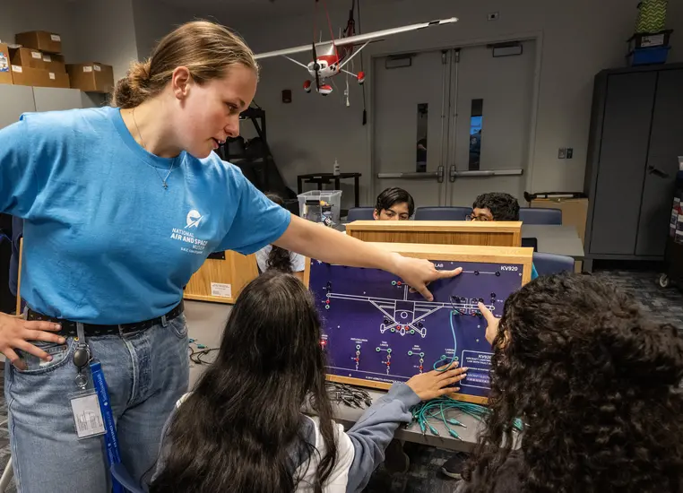 A young woman in a camp t-shirt points to a point on a board with a model of an airplane, as campers work on the plane. 