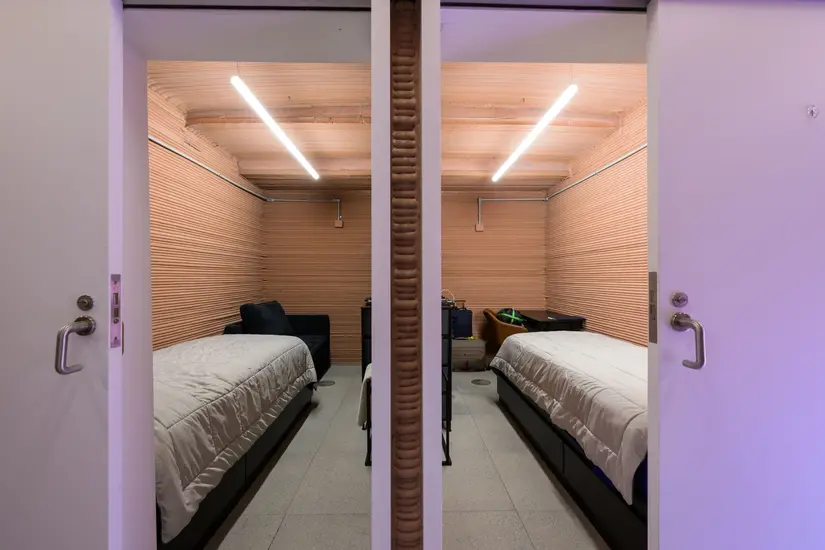 A photo looking into two rooms, with a wall separating them in the middle of the shot. In each room is a bed and a chair.