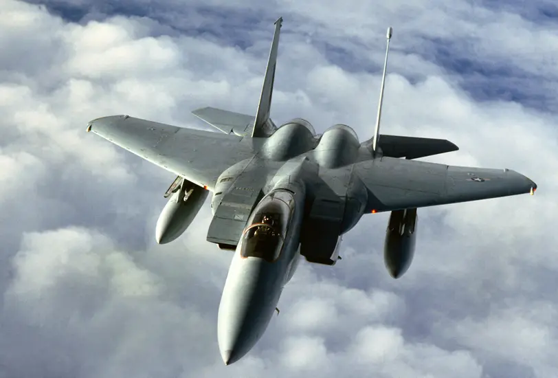 An F-15 fighter jet flying through the sky with clouds in the background.
