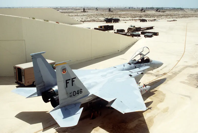 A U.S. Air Force F-15 Eagle fighter jet parked on a tarmac, with various military vehicles and equipment visible in the background.