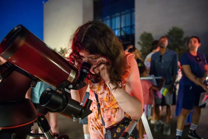 A young girl looks through the lens of a red telescope that is pointed at the night sky. The National Air and Space Museum is in the background. 